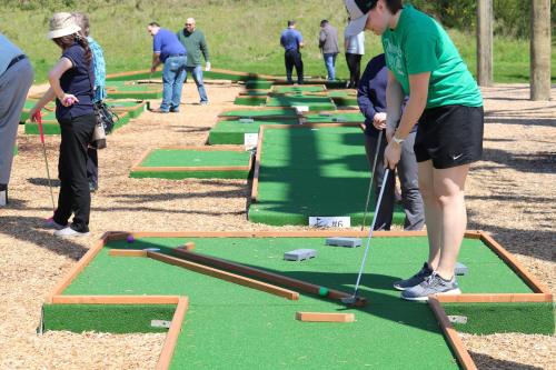 A woman in a green shirt prepares to putt on a mini-golf course, with other players in the background.