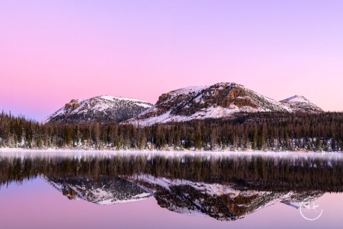 Serene landscape of snow-capped mountains reflecting in a calm lake at dawn, with a pastel sky above.