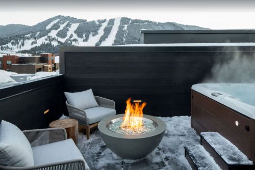 A cozy rooftop terrace with a fire pit, seating, and a hot tub, overlooking snow-covered mountains.