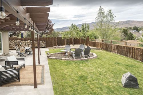 A spacious backyard with a stone fire pit, seating area, and lush green grass, surrounded by trees and mountains.