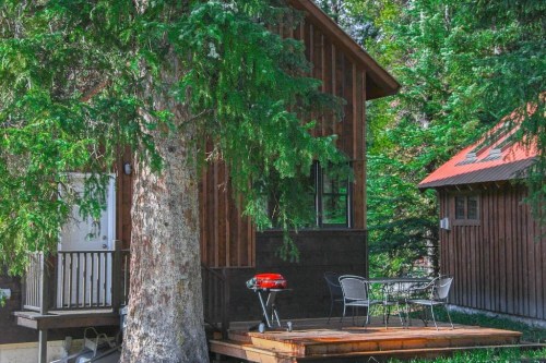 A wooden cabin surrounded by trees, featuring a small deck with a grill and outdoor seating.
