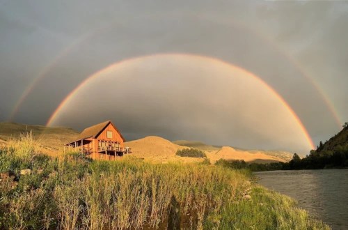 A wooden cabin by a river under a double rainbow, with rolling hills in the background and a cloudy sky.