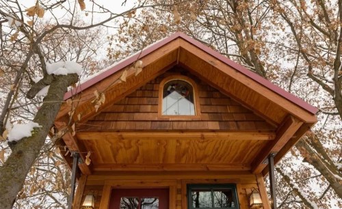 A close-up of a wooden cabin's roof with a small arched window, surrounded by snow-covered trees.