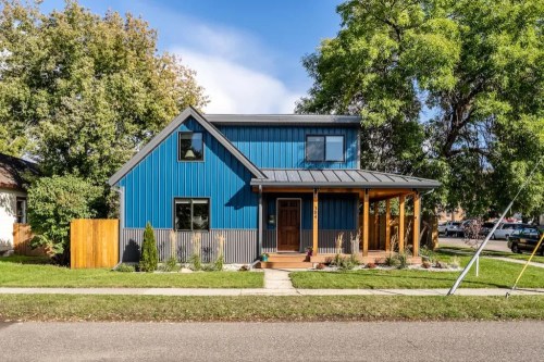 A modern blue house with a metal roof, front porch, and landscaped yard on a sunny street.