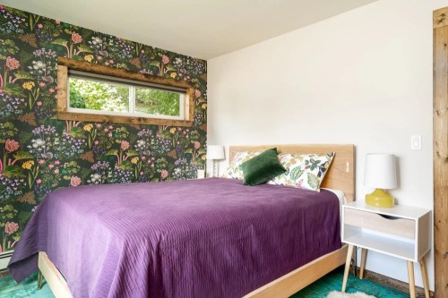 Cozy bedroom with floral wallpaper, a purple bedspread, and a wooden bed frame, featuring a nightstand and green accents.