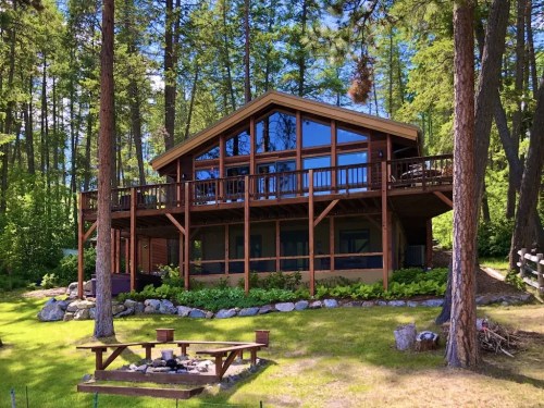 A modern cabin surrounded by trees, featuring large windows and a deck, with a fire pit in the foreground.