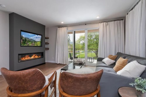Modern living room with a gray wall, TV, fireplace, large windows, and cozy seating. Bright and inviting atmosphere.