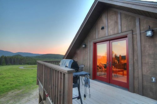 A wooden deck with a grill overlooks a scenic landscape at sunset, featuring mountains and a winding path.