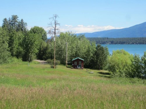 A small red cabin sits near a lake, surrounded by lush greenery and mountains under a clear blue sky.