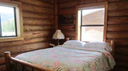 Cozy log cabin bedroom with a wooden bed, floral bedding, and a lamp by the window. Natural light fills the room.
