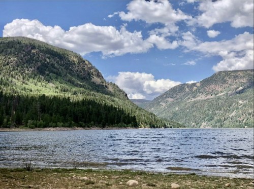 A serene lake surrounded by lush green mountains and a partly cloudy blue sky.