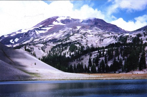 A serene mountain landscape with snow-capped peaks, lush green trees, and a calm lake reflecting the sky.