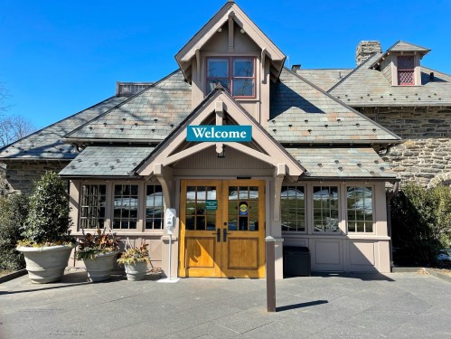 A charming building with a "Welcome" sign, featuring a sloped roof and large windows, surrounded by potted plants.