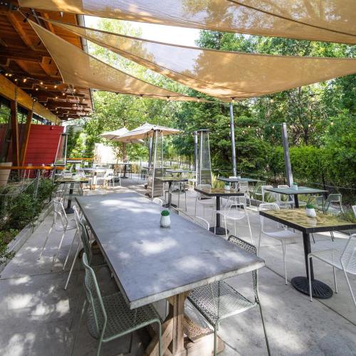 Outdoor dining area with tables and chairs under shade sails, surrounded by greenery and a restaurant in the background.