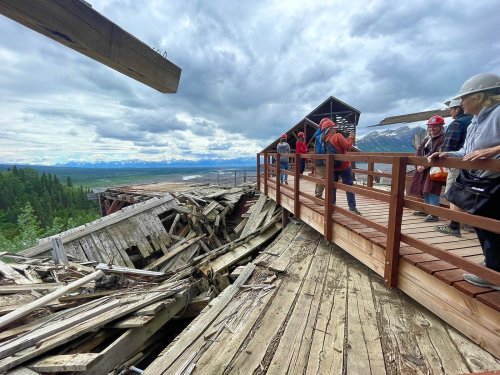 A group of people on a wooden walkway overlooking a damaged structure and a scenic landscape under cloudy skies.
