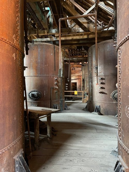 Interior of an industrial space featuring large, rusty tanks and wooden flooring, with beams and pipes overhead.