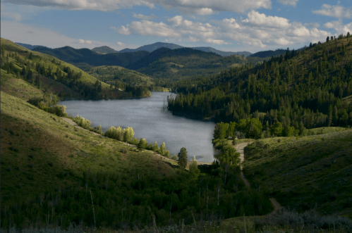 A serene landscape featuring a lake surrounded by lush green hills and trees under a partly cloudy sky.