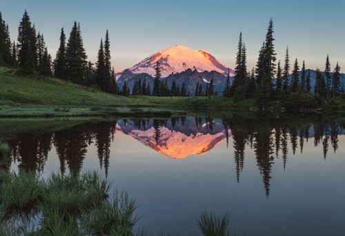 A serene landscape featuring a mountain reflecting in a calm lake, surrounded by evergreen trees at sunset.