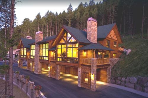 A large, modern log cabin with stone accents, surrounded by trees, illuminated at dusk.