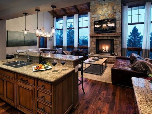 Cozy living room with a stone fireplace, large windows, and a kitchen island featuring snacks and wine. Warm, inviting atmosphere.