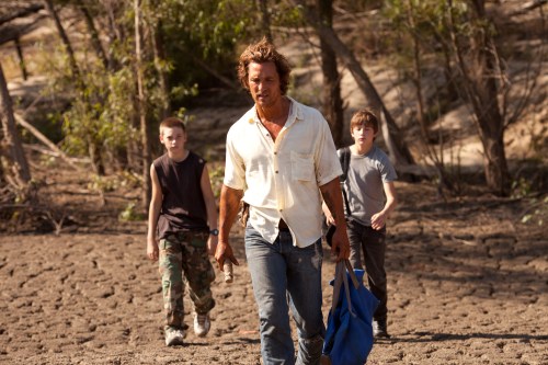A man walks on dry ground with two boys, surrounded by trees, suggesting a rural or wilderness setting.