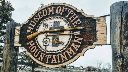 Sign for the Museum of the Mountain Man featuring a rifle and decorative elements, set against a natural backdrop.