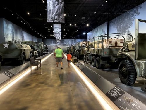A man and child walk through a military museum, surrounded by vintage vehicles and historical displays.