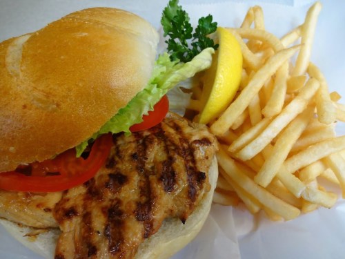 A grilled chicken sandwich with lettuce, tomato, and a lemon wedge, served with a side of crispy French fries.