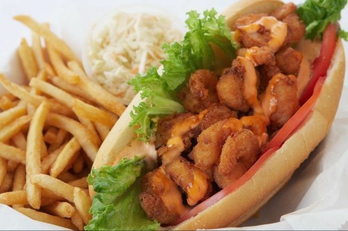 A shrimp po' boy sandwich with lettuce, tomato, and spicy sauce, served with fries and a side of coleslaw.