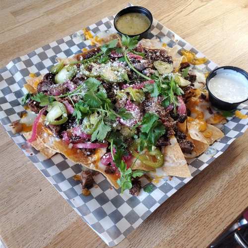 A plate of loaded nachos topped with meat, cheese, pickled onions, jalapeños, and fresh cilantro, served with sauces.