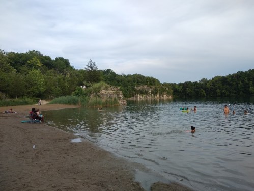 A sandy beach by a calm lake with people swimming and relaxing, surrounded by greenery and rocky cliffs.