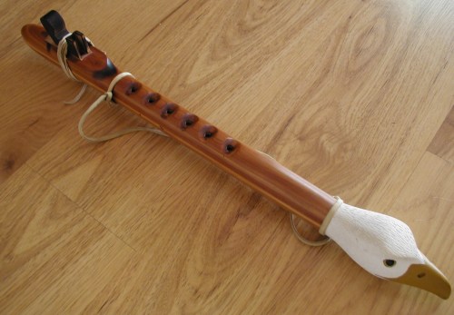 A wooden flute with a carved bird head, featuring finger holes and decorative leather ties, resting on a wooden surface.