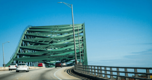 A green bridge with a unique design rises over a roadway, with cars driving on the road beneath a clear blue sky.