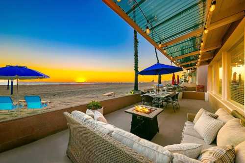 Cozy beachside patio with a fire pit, seating, and umbrellas, overlooking a vibrant sunset over the ocean.