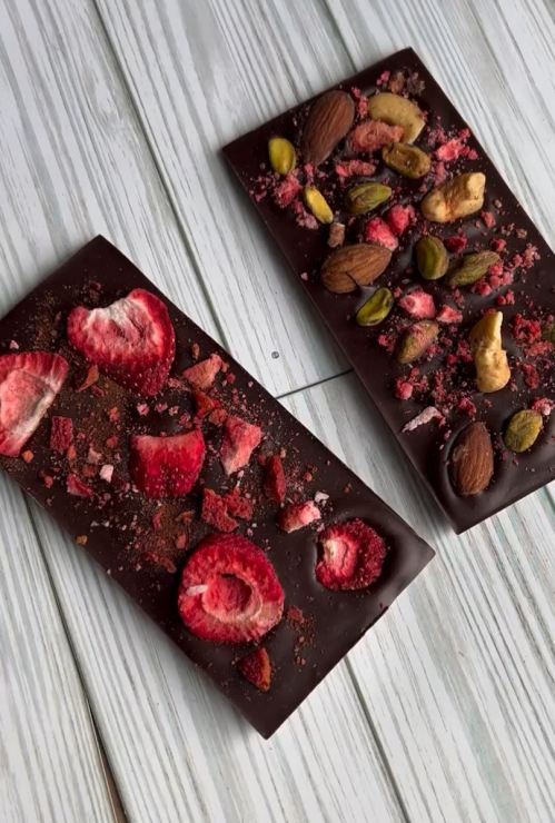 Two chocolate bars topped with dried strawberries, nuts, and crushed fruit, resting on a light wooden surface.