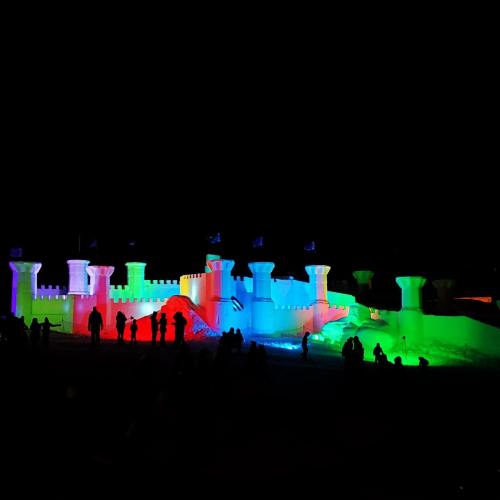 A brightly lit ice castle at night, featuring colorful lights and silhouettes of people in front.