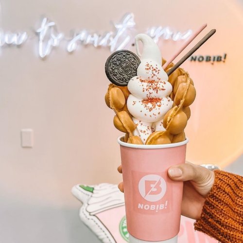 A hand holding a pink cup filled with soft serve ice cream, topped with an Oreo and waffle balls, against a light background.