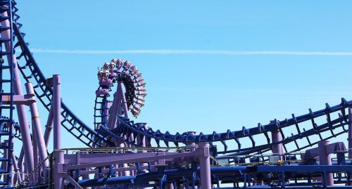 A purple roller coaster twists against a clear blue sky, with riders at the top of a loop.