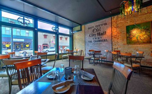 Interior of a restaurant with wooden tables and chairs, featuring a brick wall and large windows overlooking the street.
