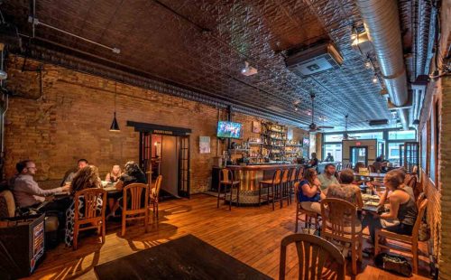 A cozy restaurant interior with wooden floors, a bar, and patrons enjoying meals at tables. Warm lighting creates a welcoming atmosphere.