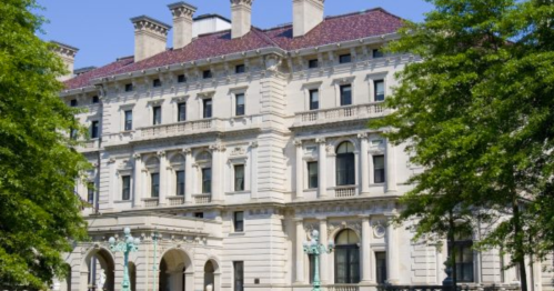 Historic mansion with ornate architecture, surrounded by lush green trees and a clear blue sky.