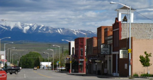 A small town street lined with historic buildings, with snow-capped mountains in the background under a blue sky.