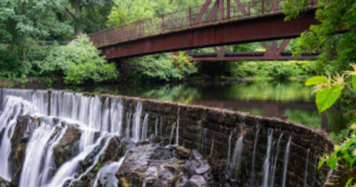 A serene waterfall flows beneath a rusted bridge, surrounded by lush greenery and calm waters.