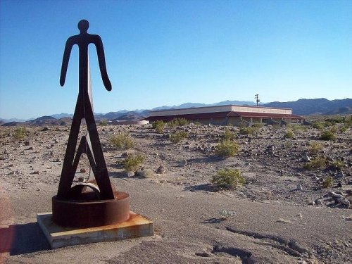 A tall metal sculpture stands in a barren landscape with mountains in the background and a building nearby.