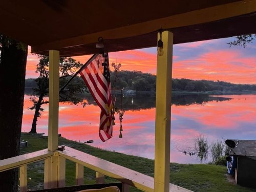 A serene lake at sunset, reflecting vibrant pink and orange hues, with an American flag hanging nearby.