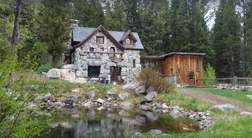 A charming stone cottage with a wooden addition, surrounded by greenery and a small pond, set in a forested area.