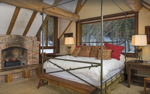 Cozy bedroom with a four-poster bed, decorative pillows, a stone fireplace, and large windows overlooking a snowy landscape.
