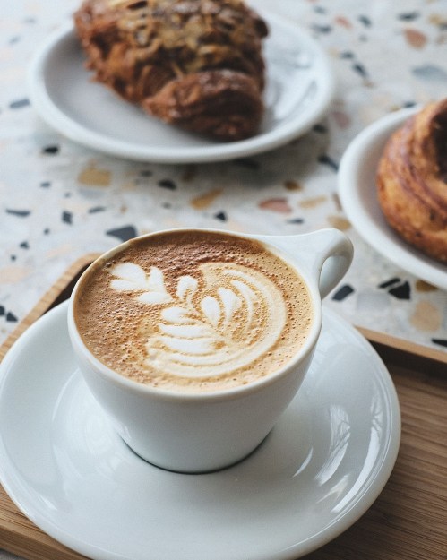 A cup of latte art on a saucer, with pastries in the background on a textured table.