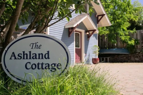 A blue cottage with a sign reading "The Ashland Cottage" surrounded by greenery and a stone pathway.