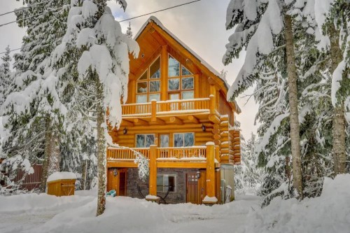 A cozy log cabin surrounded by snow-covered trees, featuring large windows and a balcony.